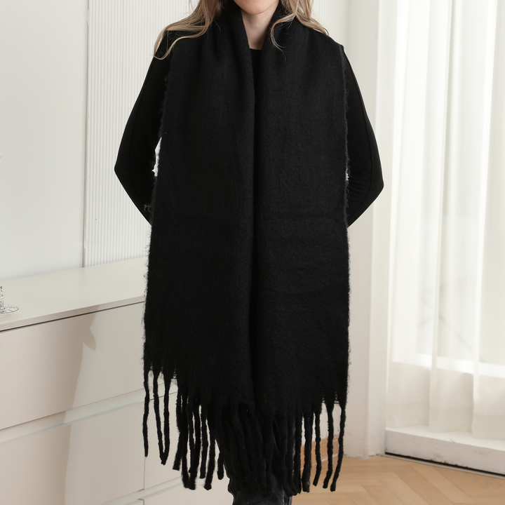 A person wearing a long black fuzzy wrap or scarf with fringe along the bottom, photographed indoors against a bright white wall and window curtains.