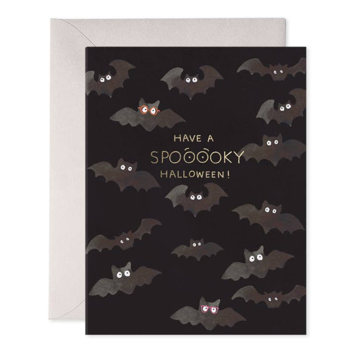 A Halloween greeting card with a black background, illustrated bats with white eyes and a few with red-pink eyes, plus the text 'HAVE A SPOOKY HALLOWEEN!' in gold. A light beige envelope is placed behind the card on a white background.