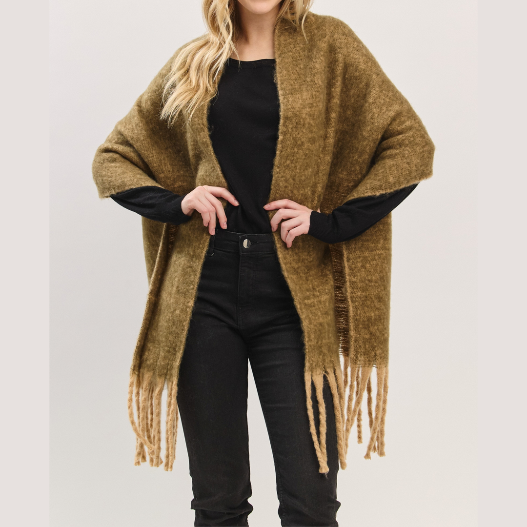 A woman wearing a chunky, open-front poncho cardigan in a warm olive-brown color with long fringe tassels, styled over a black top and black jeans against a light gray studio background.