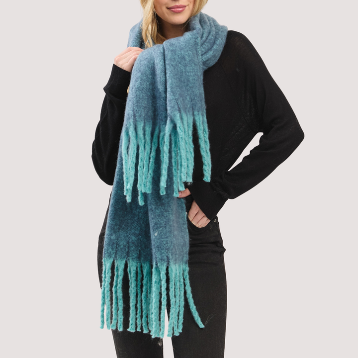 A woman wearing a fuzzy knit scarf with a blue-to-teal gradient and fringe ends, styled over a black sweater against a light gray studio background.