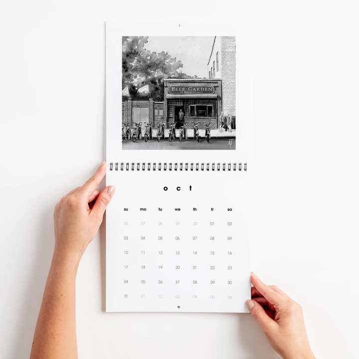 A spiral-bound wall calendar held open by two hands against a plain white wall. The top page shows a black-and-white photograph of a storefront with the words "Cafe Cuisinier," and the bottom page displays the month grid for October.