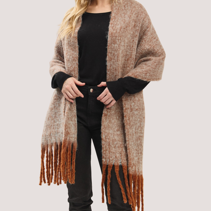 A woman wearing a long, open-front fuzzy knit poncho in a heathered taupe-brown color with darker brown fringe tassels at the bottom, styled over a black top and black pants against a light gray studio background.