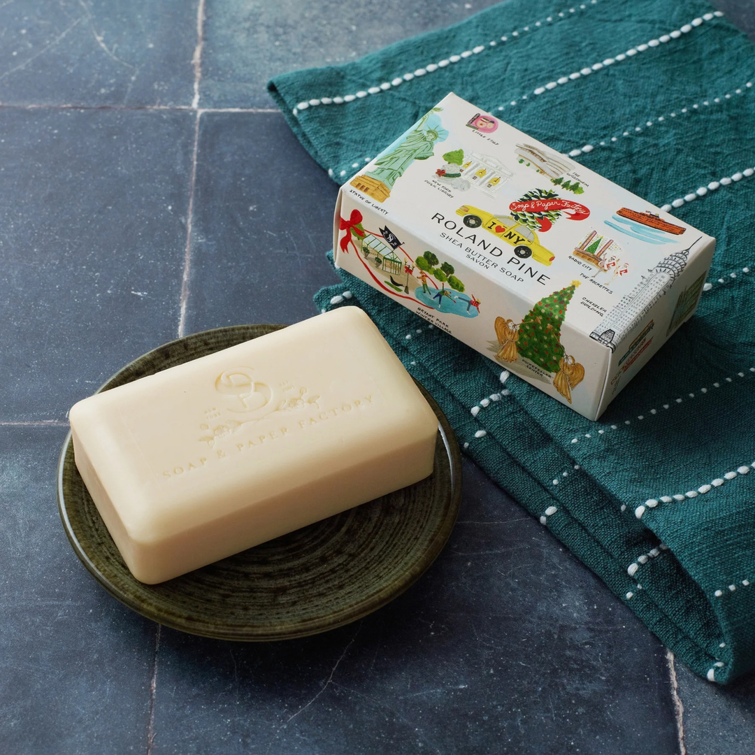 A bar of soap resting on a small dark plate next to its illustrated retail box, placed on a teal patterned towel over a dark stone surface.