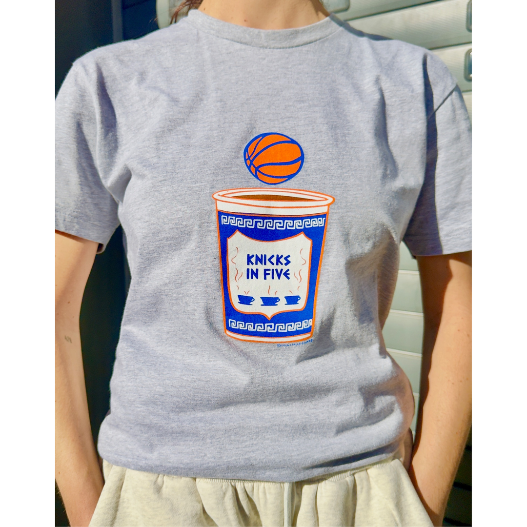 Woman standing in gray tshirt that features an anthora cup that says "knicks in five" with a basketball above it.