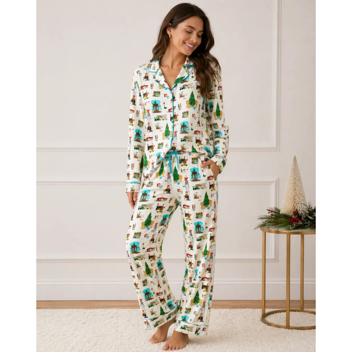 A woman stands barefoot in a bedroom wearing a matching long-sleeve pajama set with a holiday village print on an off-white background. The print features green trees, red houses, and blue elements.