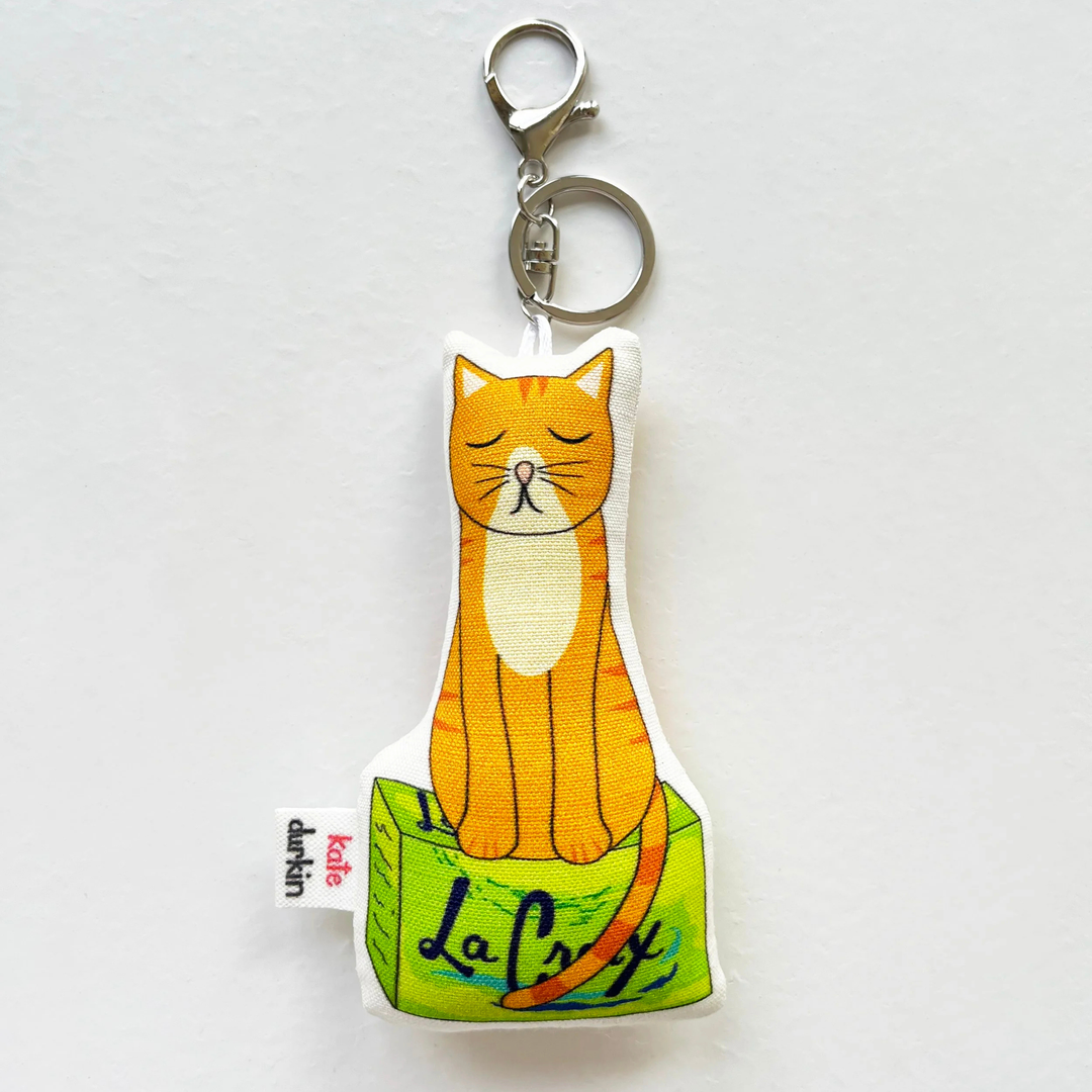 A fabric keychain charm shaped like an orange tabby cat sitting on a green box labeled &