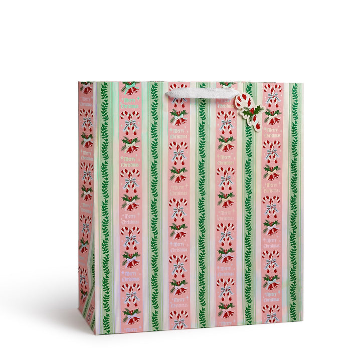 A Christmas-themed gift bag with a vertical striped pattern featuring pink candy cane motifs, green holly leaves, and red berries, complete with a white handle and a hanging ornament.