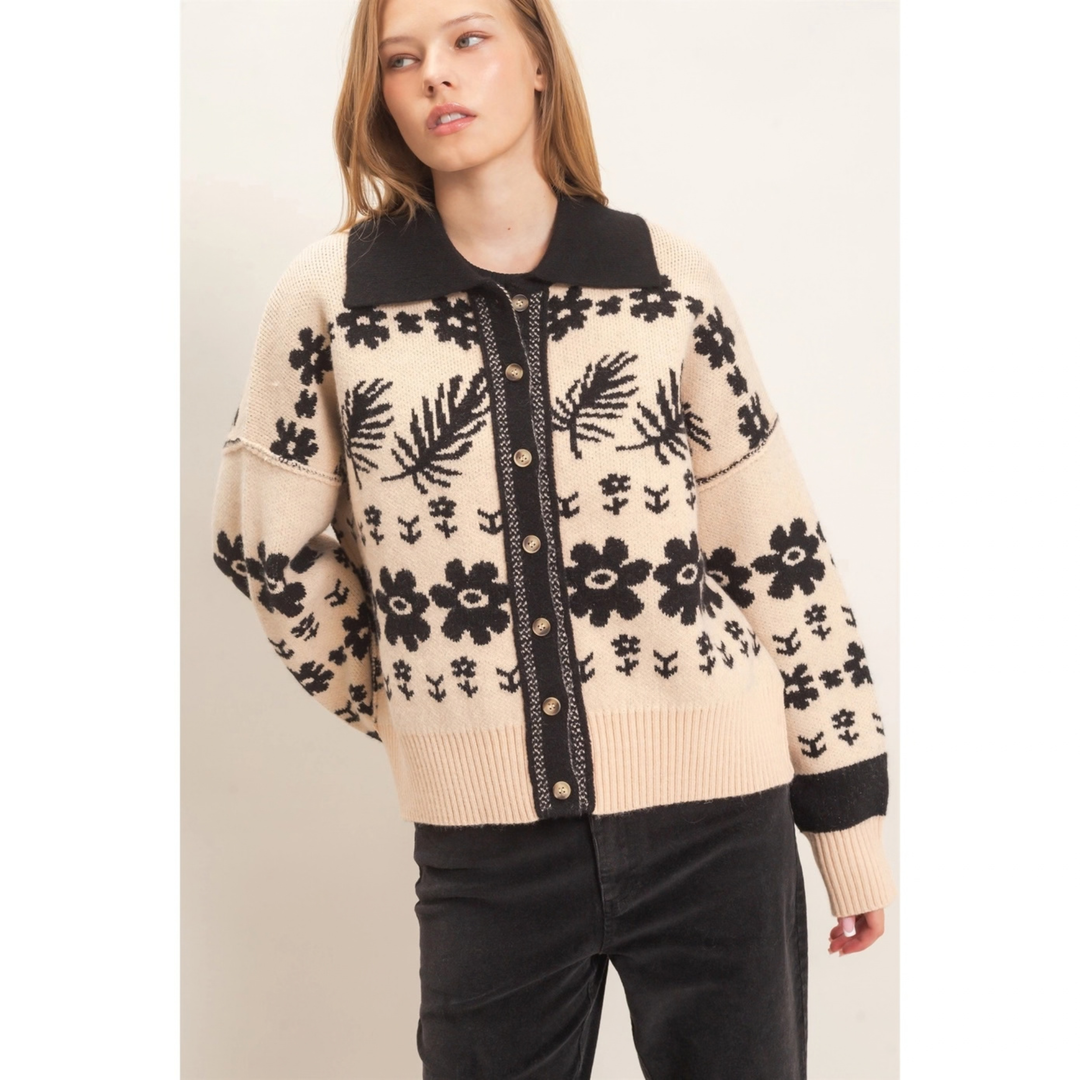 A woman wearing a cream and black knit cardigan sweater with a floral pattern, black collar and button placket, paired with black pants, against a plain light beige background.