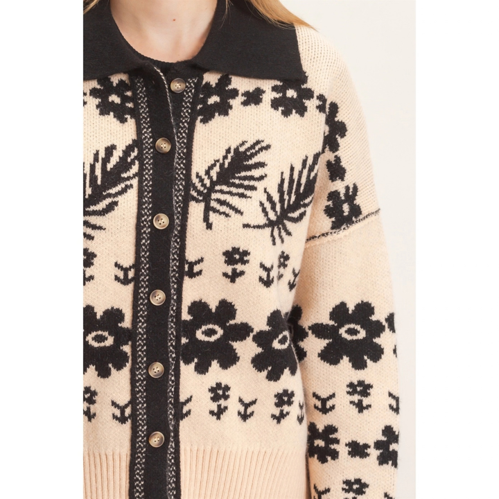 A close-up of a cream-colored knit cardigan sweater with a black collar, black leaf and flower patterns, and a black button placket with gold-tone buttons.