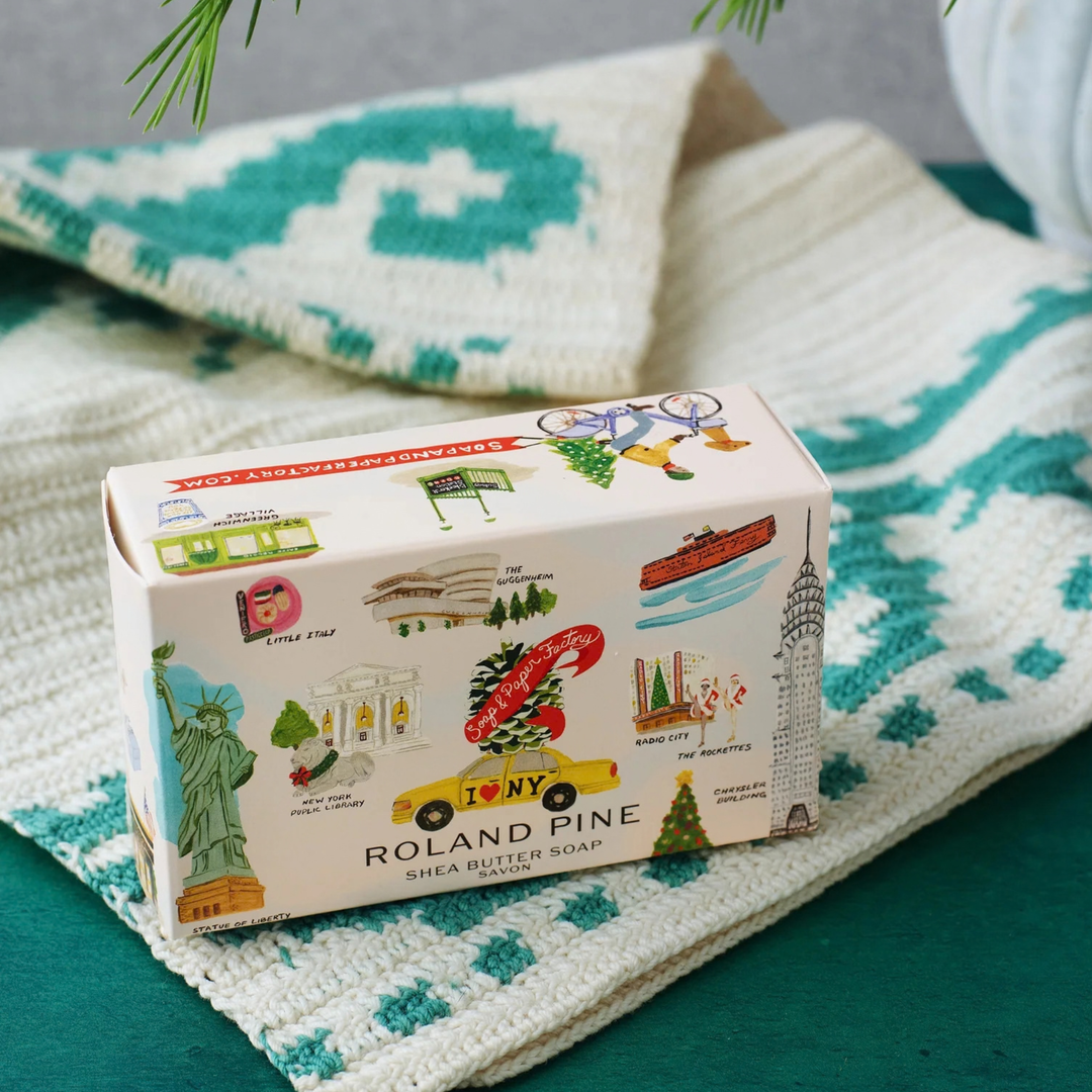 Roland Pine Shea Butter Soap bar in a decorative illustrated box, styled on a teal and white patterned knit blanket.