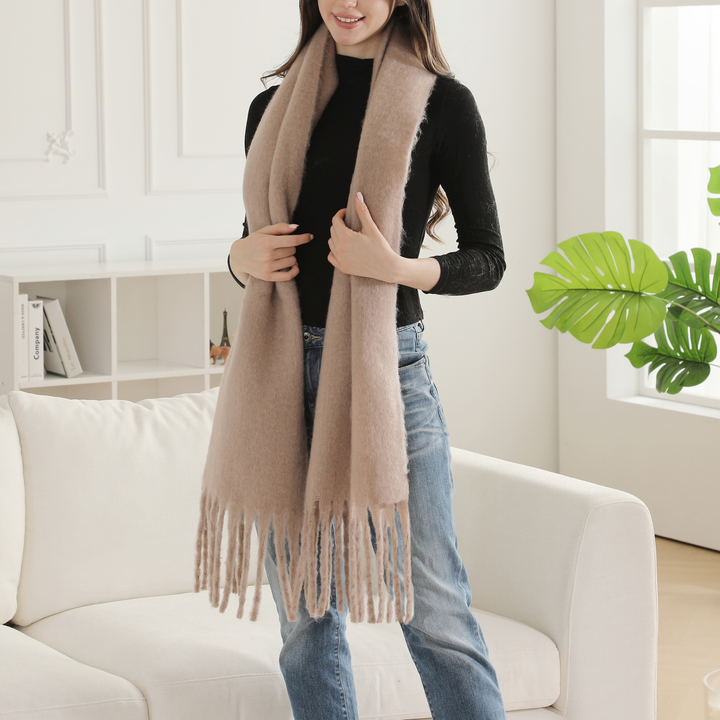 A woman wearing a long, soft beige scarf with fringe, styled over a black top and blue jeans, standing in a bright, modern living room next to a white sofa and a green plant.