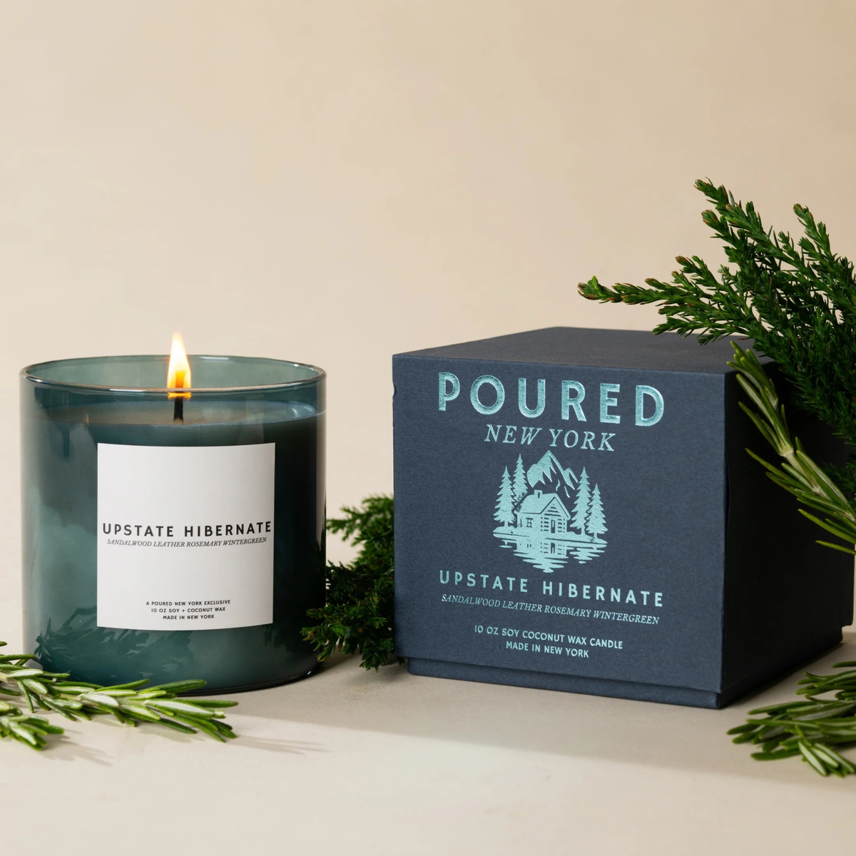 A lit teal glass candle sits beside its dark navy branded box on a light beige surface, styled with green pine branches. The label reads UPSTATE HIBERNATE, and the box shows Poured New York.