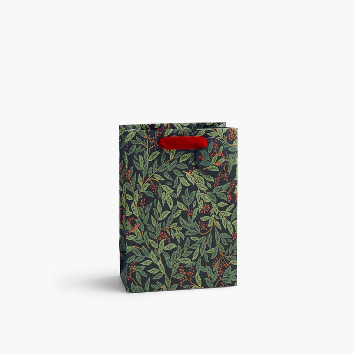 A tall paper gift bag with a dark green and black botanical pattern of leaves and berries, featuring red paper handles, set against a plain white background.