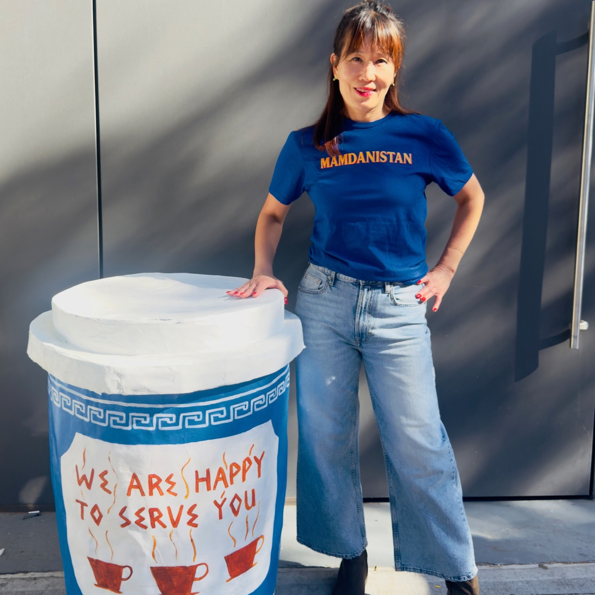 A woman stands outdoors next to a large novelty coffee cup decoration. She is wearing a blue t-shirt with 'MAMDANISTAN' printed in orange text.