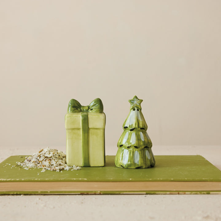 Two ceramic shakers. One is shaped as a a green Christmas tree with a gold star on top and a cream-colored gift box with a green ribbon and bow. 