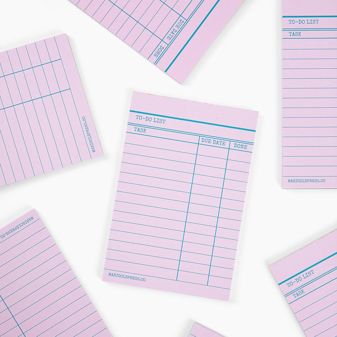 Pink lined notepad pages with blue lines and printed task columns, arranged in a flat-lay pattern on a white background.