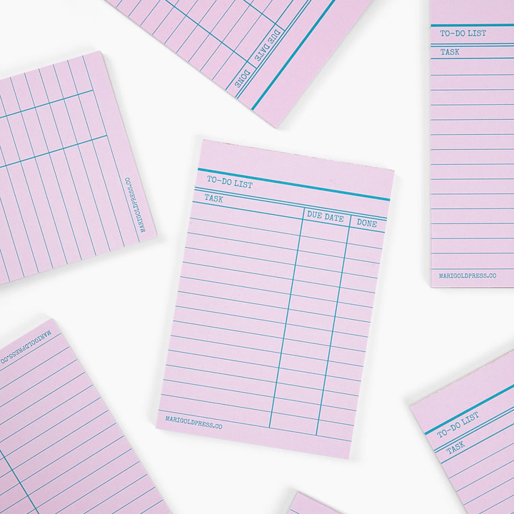 Pink lined notepad pages with blue lines and printed task columns, arranged in a flat-lay pattern on a white background.