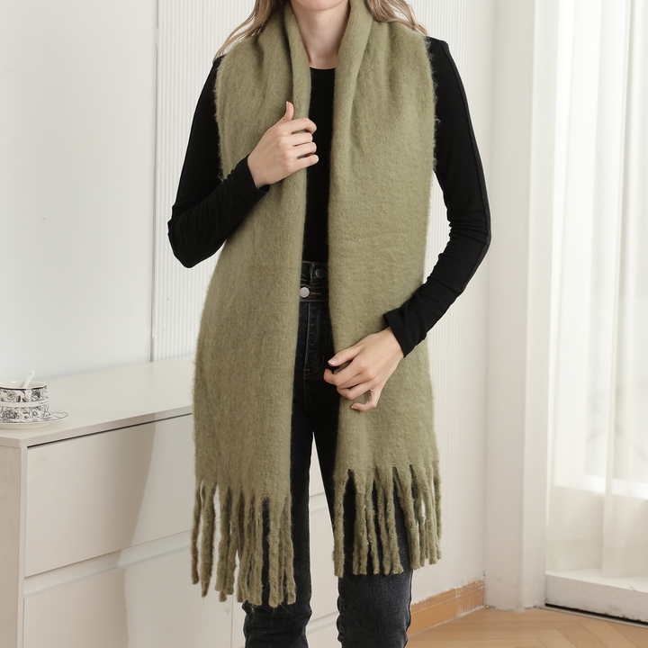 A woman wearing a long, fuzzy olive green fringed scarf over a black long-sleeve top and dark jeans, standing indoors near a white cabinet and window.