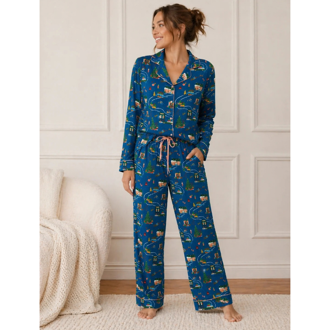A woman stands barefoot in a bedroom wearing a blue long-sleeve pajama set with an allover illustrated print of roads, cars, trees, and houses. The set includes a button-front top and matching pants with a drawstring waist.