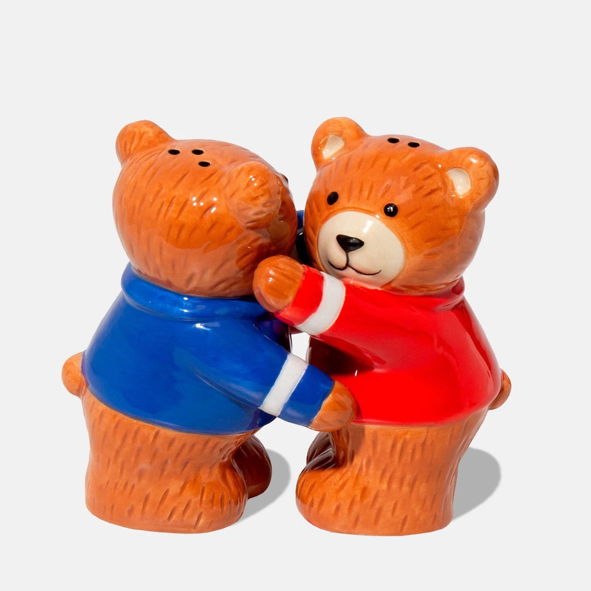 Two ceramic bear salt and pepper shakers standing together and holding each other, one wearing a blue sweater with white trim and the other wearing a red sweater with white trim, on a light gray background.