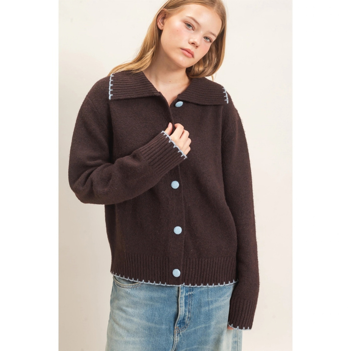 A woman wearing a dark brown knit cardigan with a wide collar, light blue buttons, and white contrast stitching, paired with light blue jeans, against a plain off-white background.
