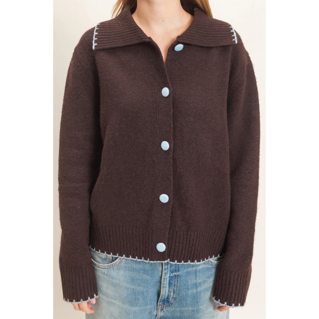 A woman wearing a dark brown knit cardigan sweater with a wide collar, light blue buttons, and white contrast stitching, paired with blue jeans.
