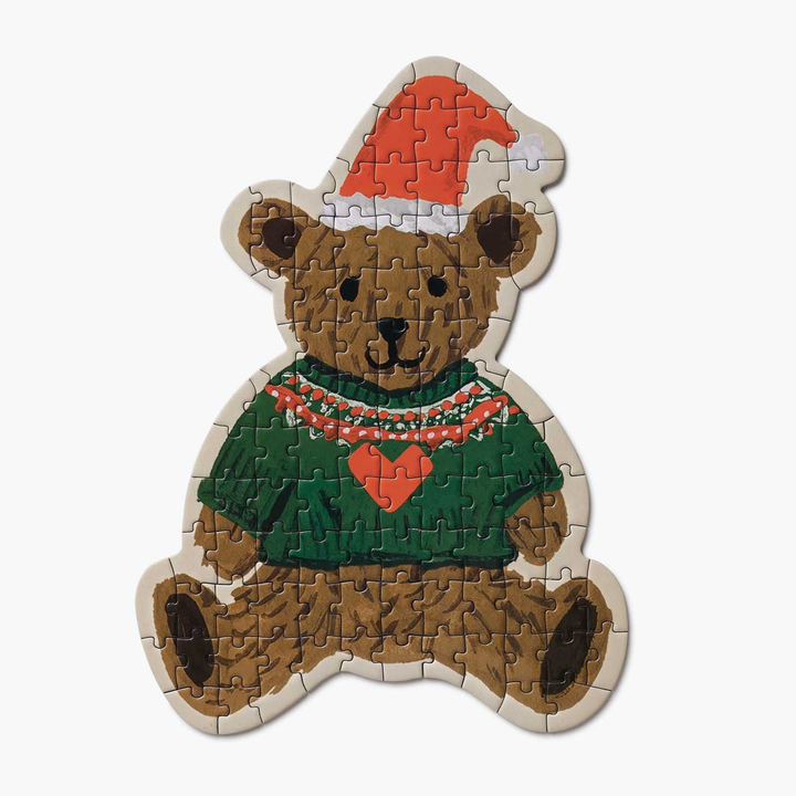 A teddy bear-shaped jigsaw puzzle featuring a brown bear wearing a red Santa hat, a green sweater with a red heart, and a red-and-white patterned scarf. The bear has dark eyes, a black nose, and a smiling expression against a plain white background.
