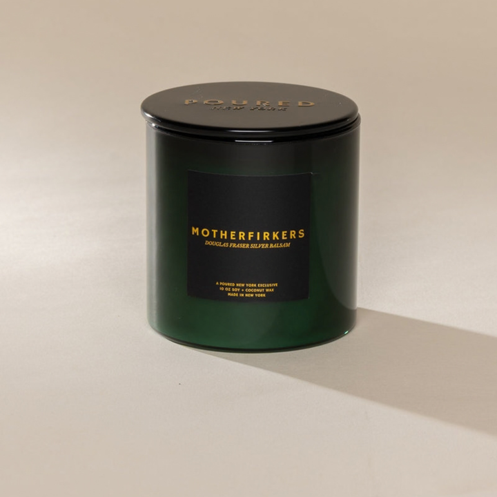 A dark green glass jar candle with a black lid and a black label featuring gold lettering that reads &