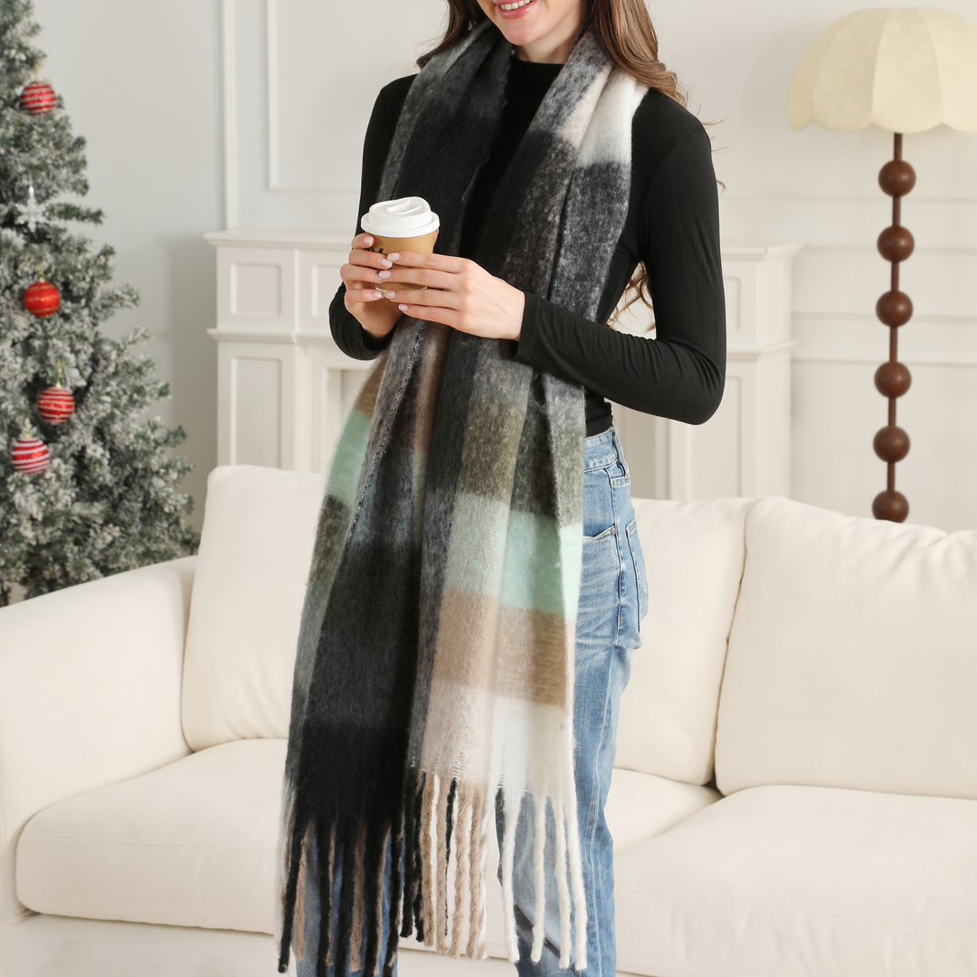 A woman wearing a long plaid scarf in black, white, beige, and light green, paired with a black top and blue jeans, holding a coffee cup in a cozy living room setting.