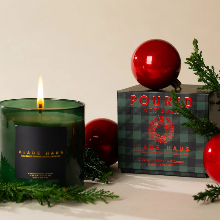 A lit Klaus Haus candle in a dark green glass jar sits beside its branded box on a light surface, styled with red Christmas ornaments and green pine branches against a warm off-white background.