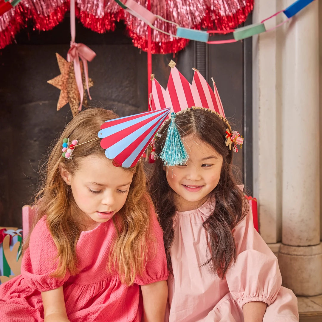 Two young girls wearing colorful party hats and hair clips, dressed in pink and coral dresses, sitting in front of a decorated fireplace with tinsel and a star ornament.