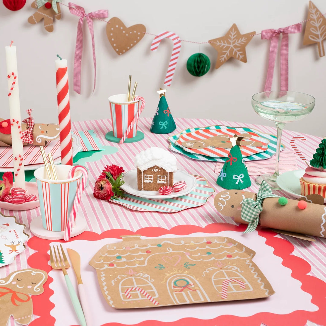 A festive Christmas gingerbread-themed party table setting with pink and white striped tablecloth, paper plates, mugs, candles, a gingerbread house placemat, and holiday decorations including a candy cane garland and paper ornaments.