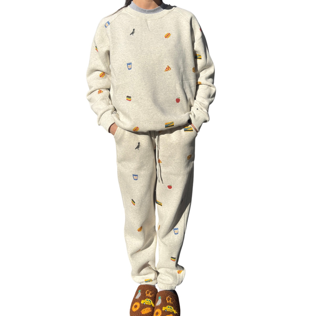 A person modeling a matching two-piece sweatshirt and jogger set in a light heather cream color, decorated with small colorful food and drink illustrations. The set includes a crewneck sweatshirt and matching drawstring pants, shown against a plain white background.