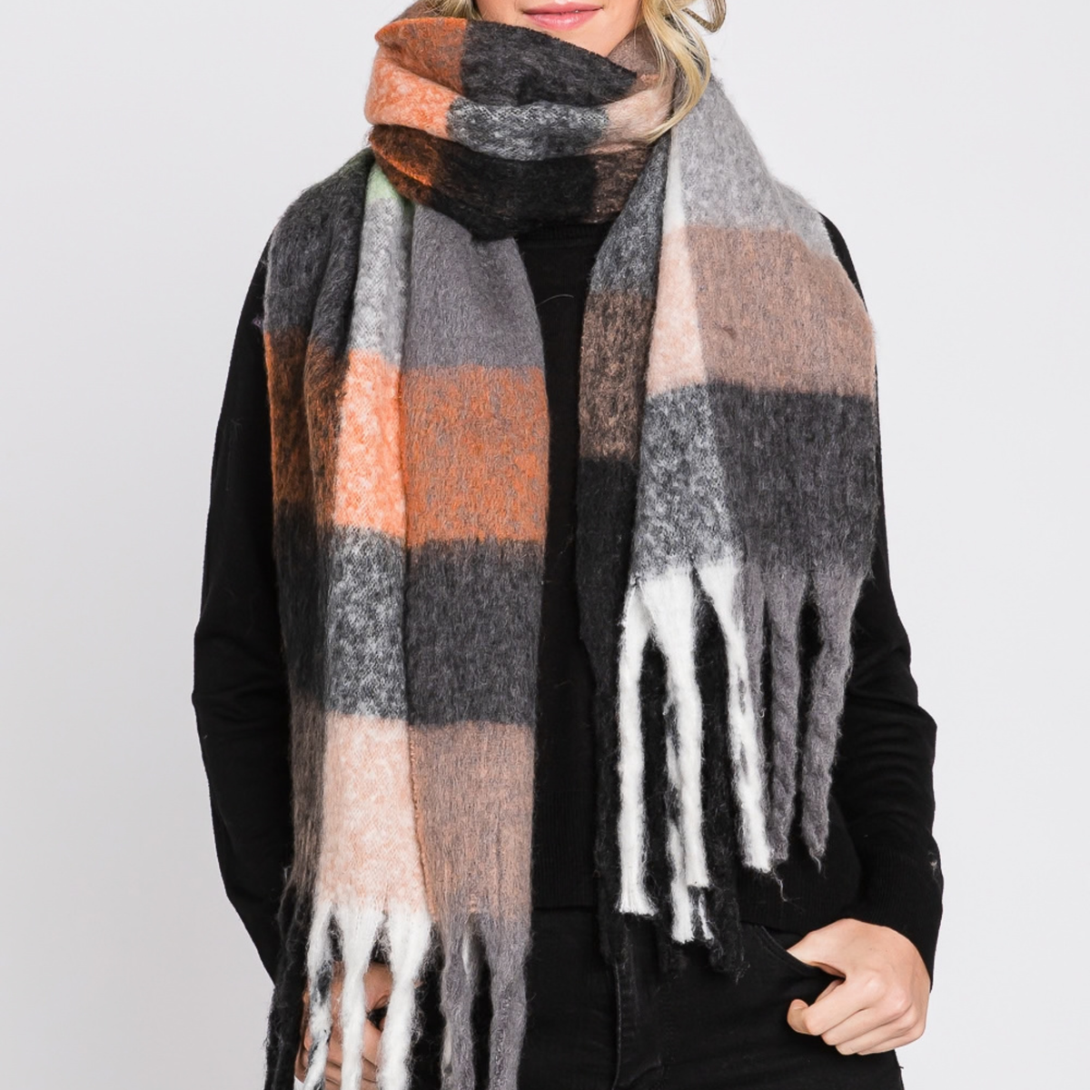 A woman wearing a thick, soft plaid scarf in shades of gray, black, peach, beige, and white, styled over a black top against a light gray background.