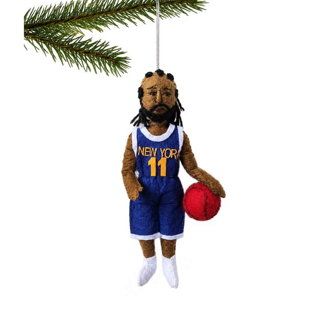 A hanging Christmas ornament shaped like a basketball player with brown skin, black dreadlocks, a beard, a blue and white jersey reading NEW YORK 11, blue shorts, white socks, and a red basketball, shown against a white background with a pine branch at the top.
