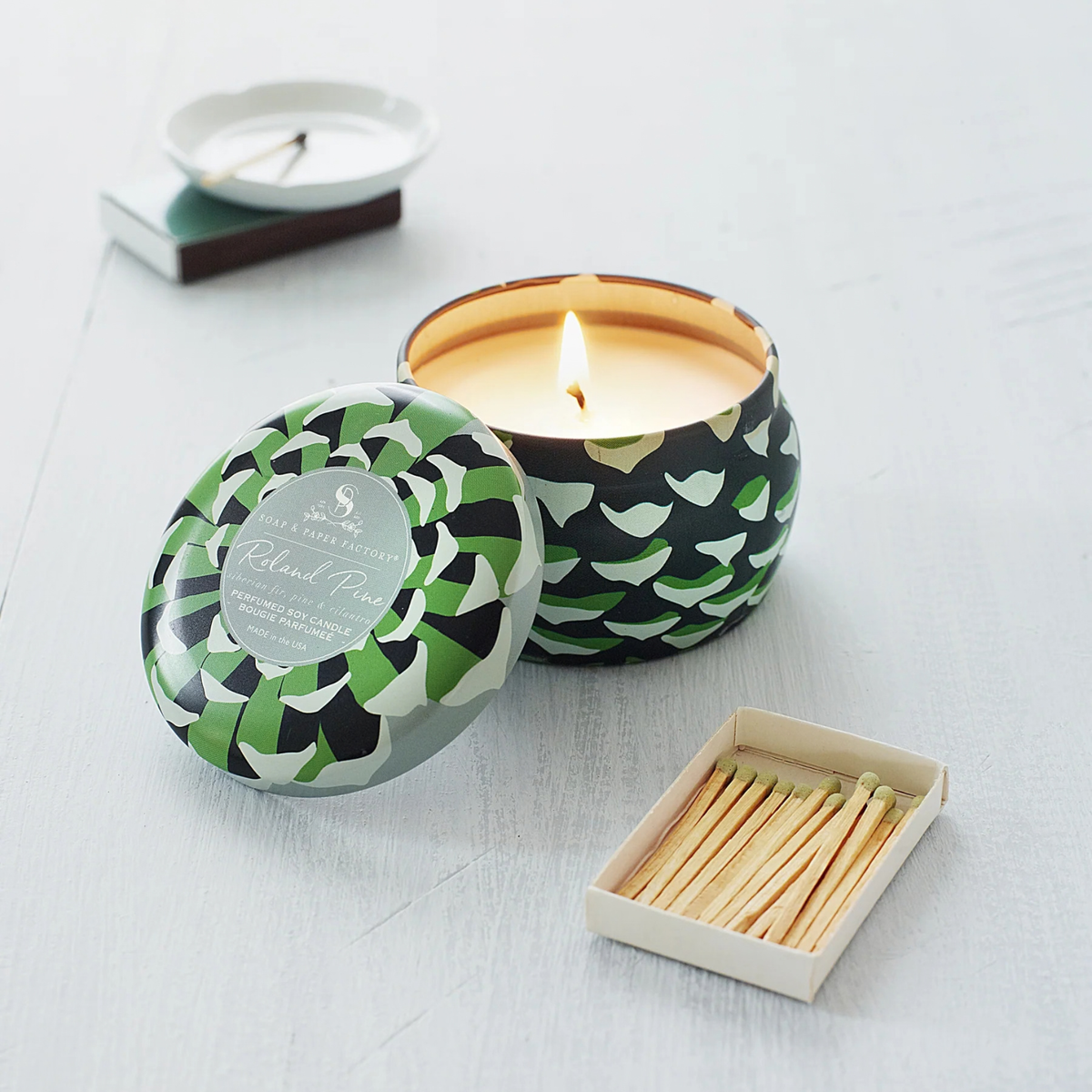 A lit candle in a patterned tin with its lid beside it, shown on a light wood surface with a matchbox and a small white dish in the background. The candle tin has a green, black, and gray geometric pattern, and the label reads 'Cloud Nine' and 'Scalpers'.