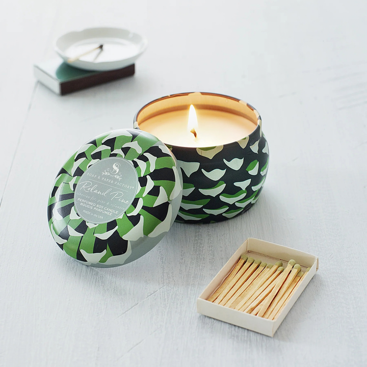 A lit candle in a patterned tin with its lid beside it, shown on a light wood surface with a matchbox and a small white dish in the background. The candle tin has a green, black, and gray geometric pattern, and the label reads &