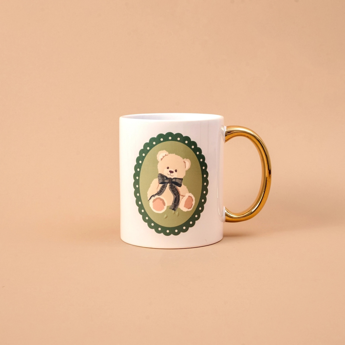 A white ceramic mug with a gold handle, featuring a printed illustration of a teddy bear in a green oval frame on the side, set against a solid beige background.