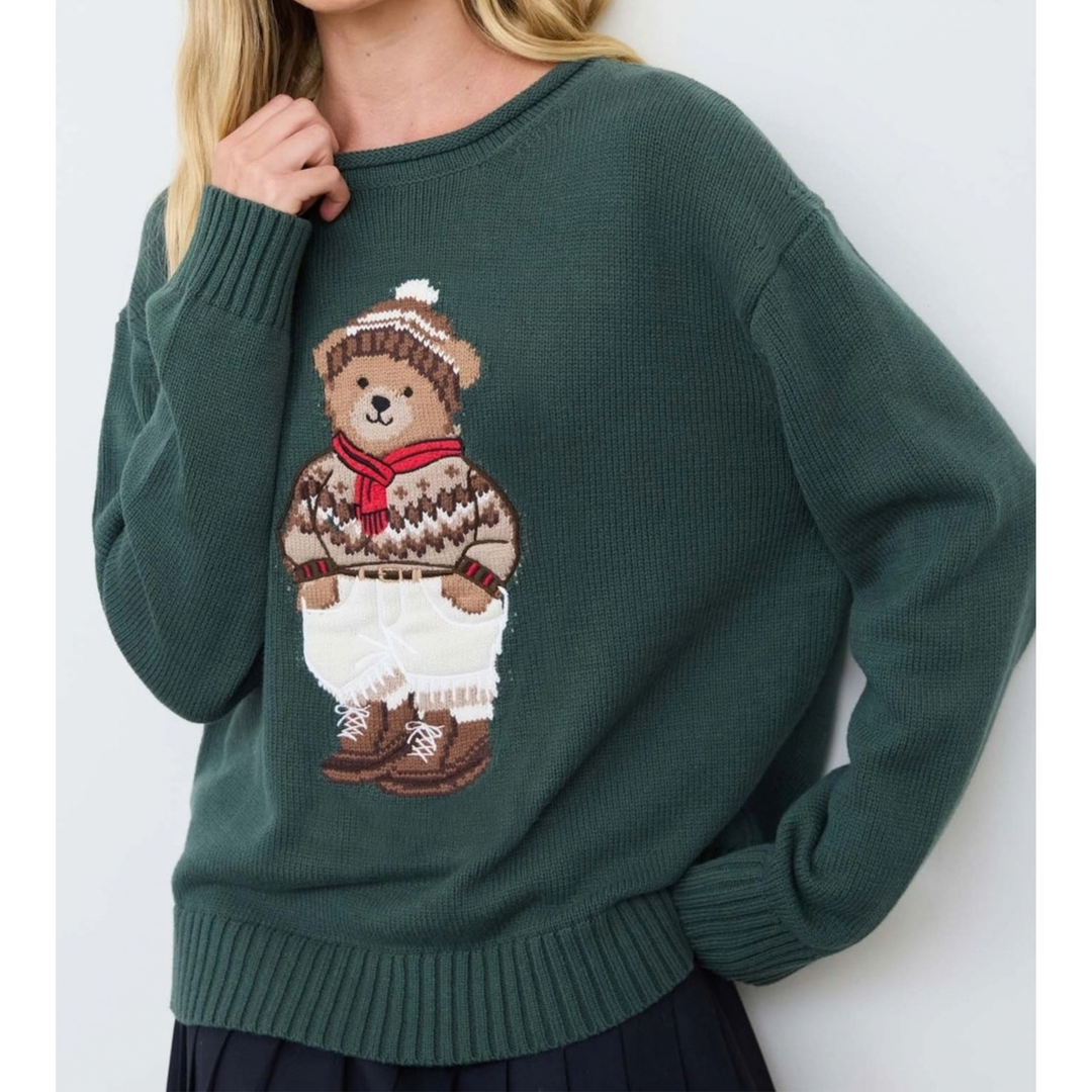 A woman wearing a dark green knit sweater featuring a large teddy bear graphic on the front, styled with a white skirt. The sweater has a ribbed crew neck, cuffs, and hem, and the bear graphic is detailed with brown, beige, and red colors.
