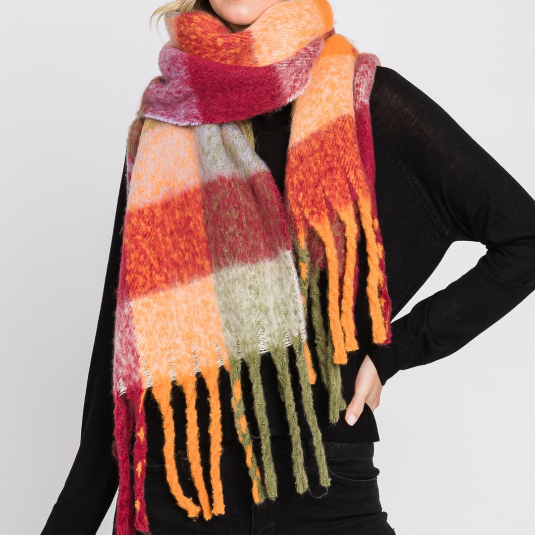A person wearing a chunky, fuzzy plaid scarf in orange, red, pink, beige, and olive green, styled over a black long-sleeve top against a light gray background.