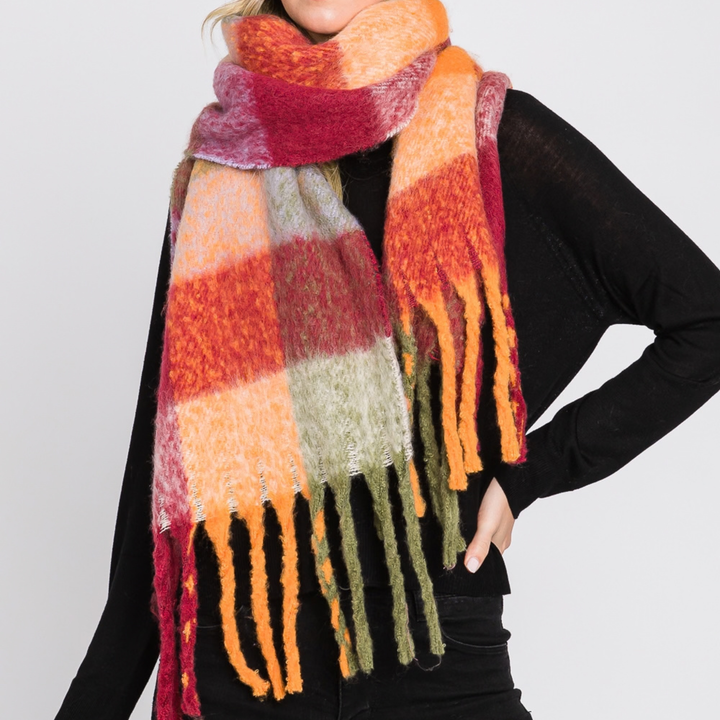 A person wearing a chunky, fuzzy plaid scarf in orange, red, pink, beige, and olive green, styled over a black long-sleeve top against a light gray background.