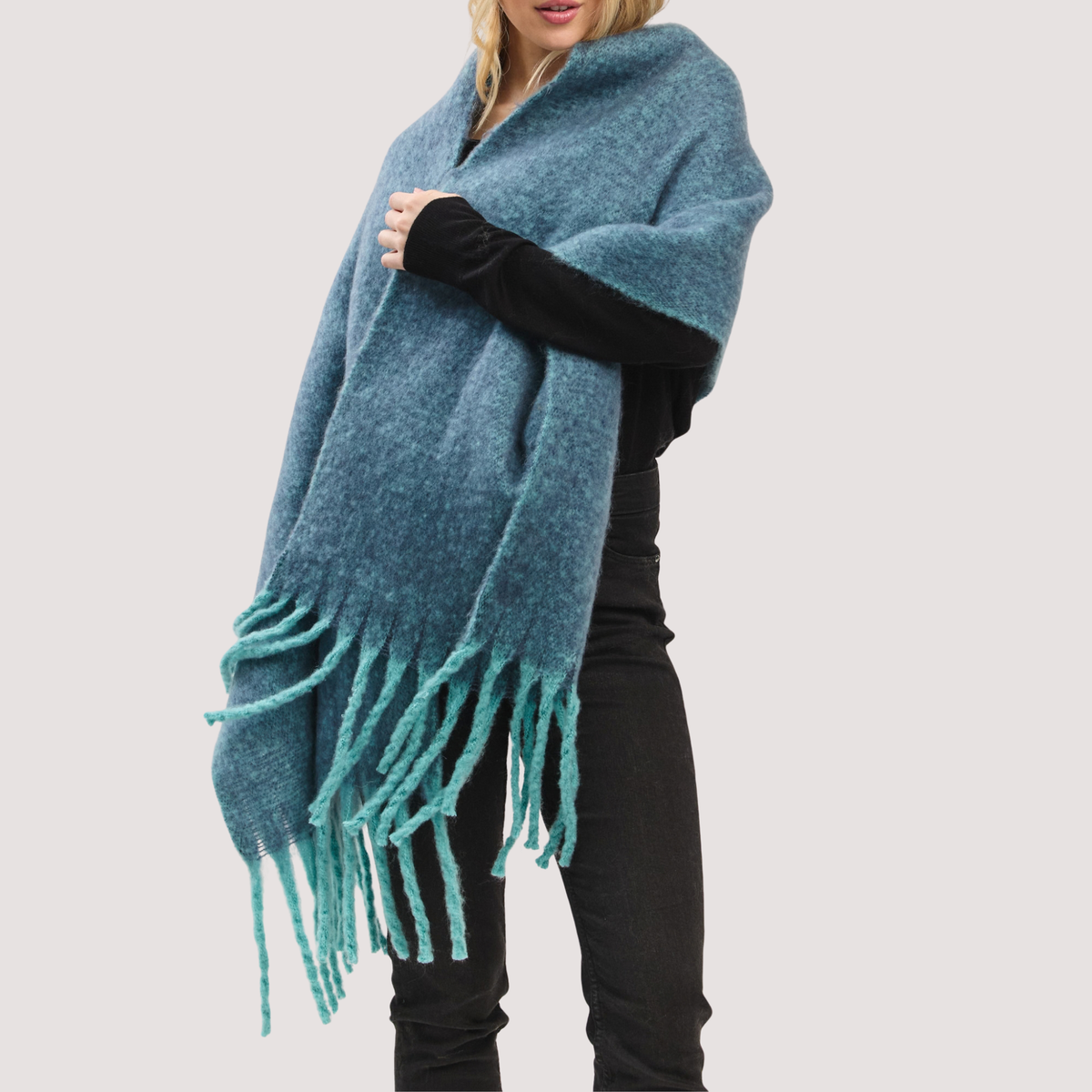A woman wearing a fuzzy teal-blue wrap poncho with long fringe, styled over a black long-sleeve top and black pants, against a plain light gray background.