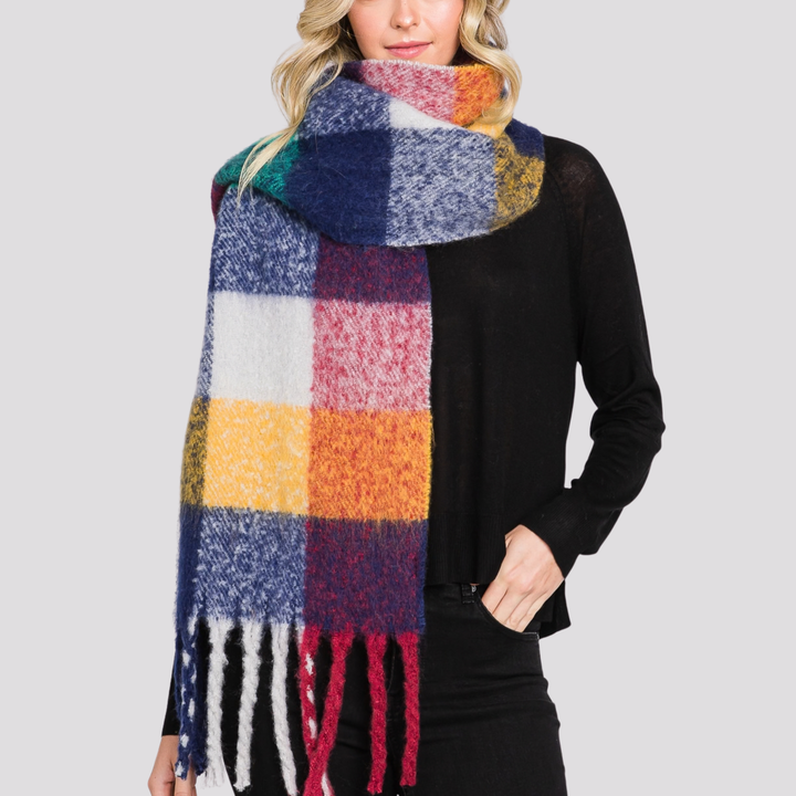 A woman wearing a chunky, multicolor plaid scarf with fringe, styled over a black long-sleeve top against a solid light gray background.