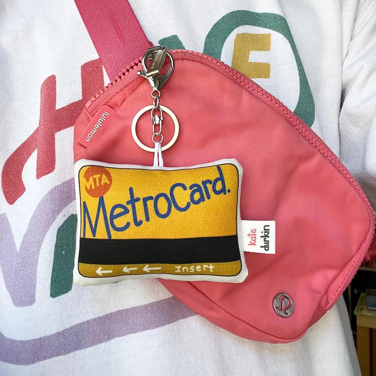 A fabric keychain designed to look like a yellow and black MetroCard with a silver metal keyring, shown clipped to a pink crossbody bag.