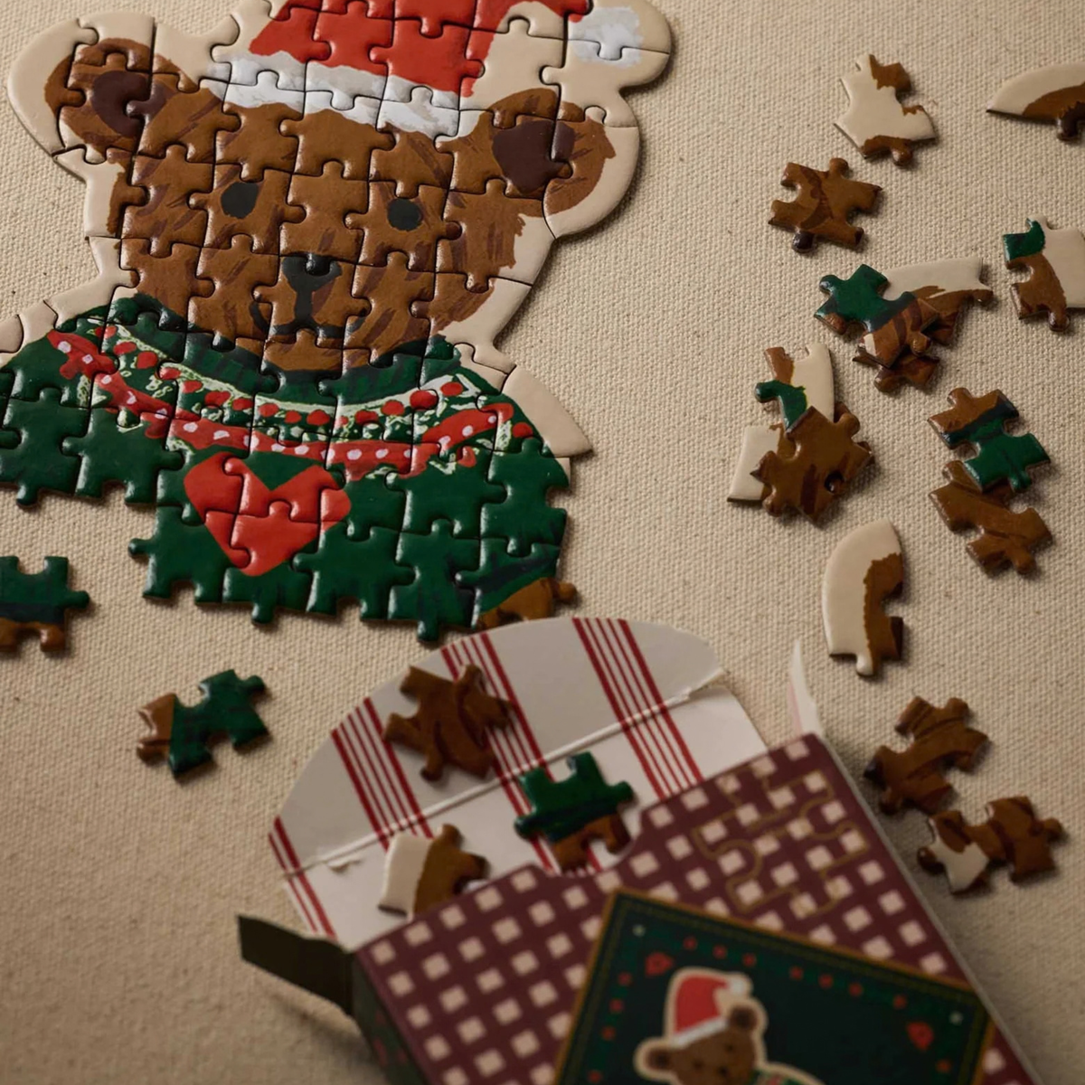A partially assembled teddy bear-shaped Christmas jigsaw puzzle on beige fabric, with the bear wearing a red Santa hat, a green sweater with a red heart, and a red-and-white scarf. Several loose brown, green, and cream puzzle pieces are scattered to the right and below the puzzle, beside an open festive gift box with red-and-white striped tissue paper and patterned Christmas packaging. Warm lighting creates a cozy holiday atmosphere.