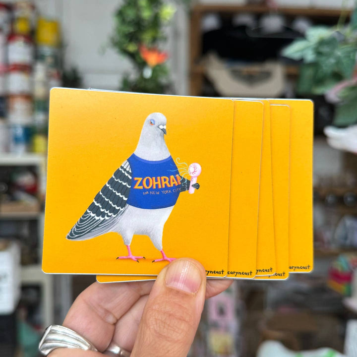 A hand holds a stack of bright yellow cards featuring an illustrated pigeon wearing a blue shirt with the word &