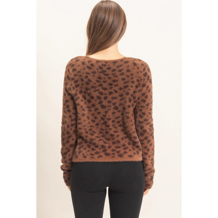 A woman viewed from the back wearing a brown fuzzy knit sweater with a dark brown leopard print pattern and black pants, set against a plain off-white background.