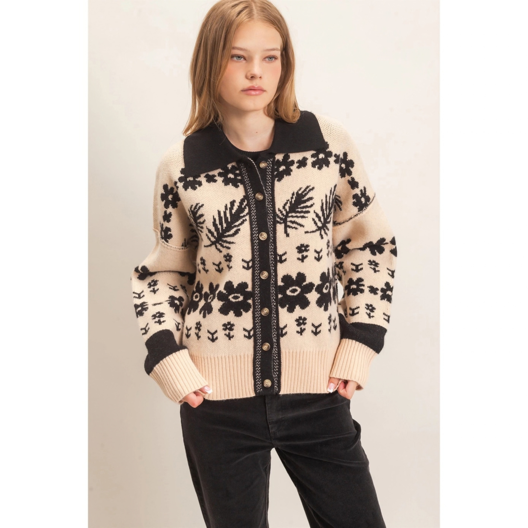 A woman modeling a cream and black knit cardigan sweater with a floral and leaf pattern, black collar and cuffs, and a button-front closure, paired with black pants against a plain light background.