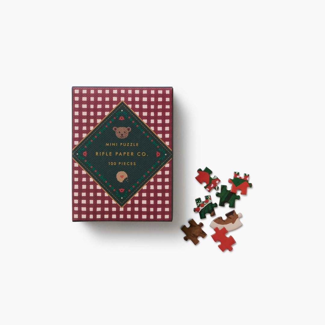 A mini Rifle Paper Co. 100-piece puzzle in a burgundy-and-cream checkered box with a dark green diamond-shaped label, gold lettering, and small animal illustrations. Several loose puzzle pieces in red, green, and brown are scattered beside the box on a white background.