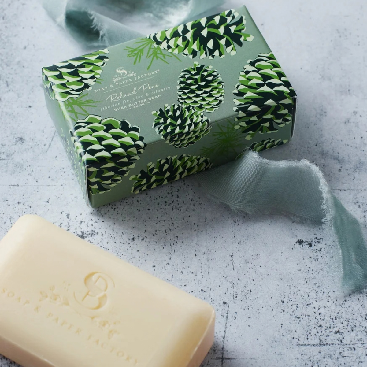 A bar of soap in a pale cream color is shown in the foreground next to its decorative packaging box, which features a green pinecone pattern on a textured gray background with a sheer ribbon.